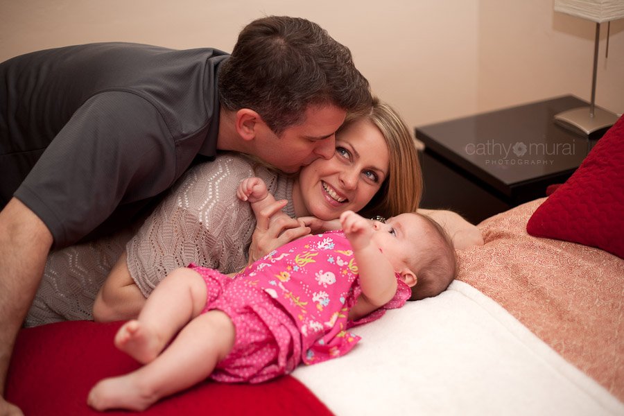 A father kissing the mom on her cheek with a beautiful baby on the bed - Captured by a Glendale Lifestyle Baby Photographer, Cathy Murai Photography
