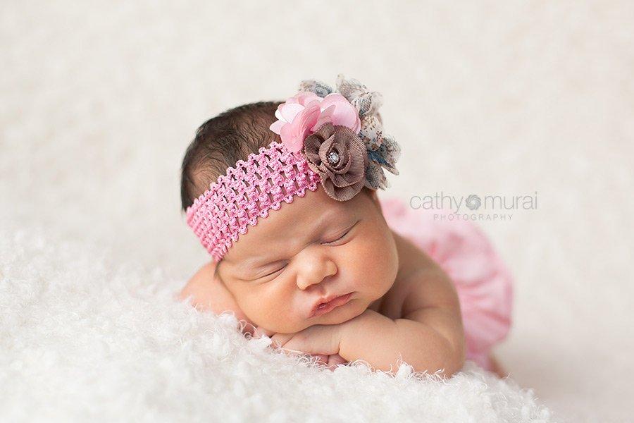 A cute newborn baby girl with chubby cheeks is wearing a pink diaper cover and a pink handmade headband.  She is posing and sleeping on the white blanket on the beach bag.  Cathy Murai Photography, Newborn and Baby Photographer in Alhambra, San Gabriel Valley, and Los angeles Area 