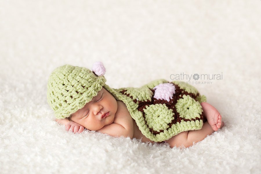 A cute newborn baby girl with chubby cheeks is wearing a handmade turtle outfit knit by her mom.  She is posing and sleeping on the white blanket on the beach bag.  Cathy Murai Photography, Newborn and Baby Photographer in Alhambra, San Gabriel Valley, and Los angeles Area 
