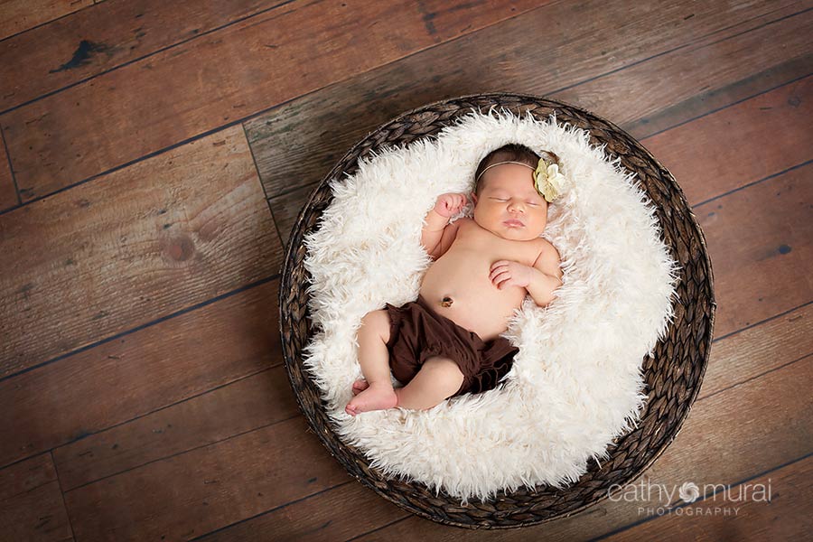 A cute newborn baby girl with chubby cheeks is wearing a brown diaper cover and cream headband.  She is posing and sleeping on the white blanket in the brown basket on the wooden floor.  Cathy Murai Photography, Newborn and Baby Photographer in Alhambra, San Gabriel Valley, and Los angeles Area 