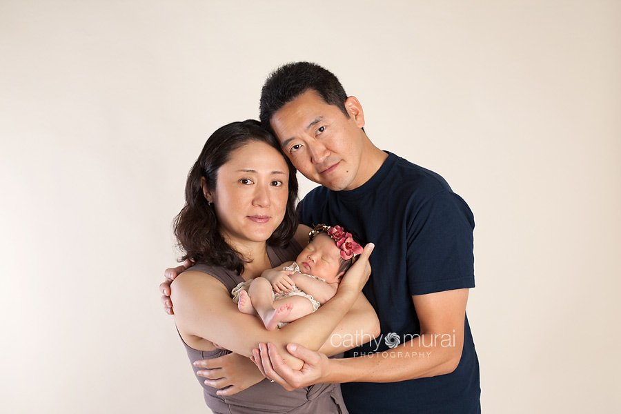 A family portrait of Asian parents and a beautiful 8 day old newborn baby girl wearing the red halo and the cream lace romper. Baby sleeping while the parents are posing in the studio for their family portrait. Los Angeles, San Gabriel Valley, Alhambra Newborn Photographer, Cathy Murai Photography