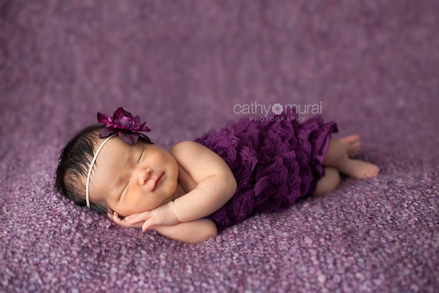 A beautiful 8 day old Asian newborn baby girl smiling wearing a purple lace romper and a purple head band on the bean bag covered with a purple blanket.  A newborn baby portrait taken in the studio during newborn portrait session by Los Angeles, San Gabriel Valley, Alhambra Newborn Photographer, Cathy Murai Photography