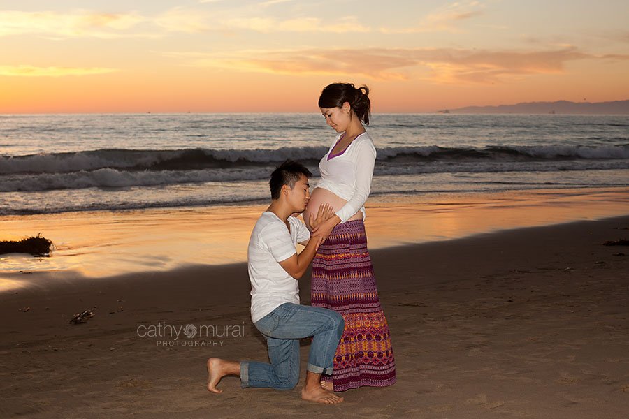 Daddy-to-be (expecting dad) is holding and kissing a gorgeous mommy-to-be's (expecting mom) belly during sunset in front of the beach at Manhattan Beach during maternity portrait session, mama-to-be, parents-to-be, expecting parents, burgundy maternity skirt, Los Angeles Maternity photographer, Manhattan Beach Session, Beach  Maternity Portrait Session, Maternity Session at Beach, Cathy Murai Photography