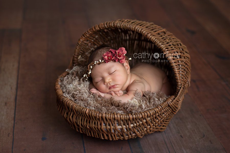 Asian newborn baby girl wearing a red flowers halo, posing and sleeping on brown furry prop inside of the brown bassinet on the wooden floor. Alhambra, San Gabriel Valley, Los Angeles, CA Studio Newborn Photographer, Cathy Murai Photography 