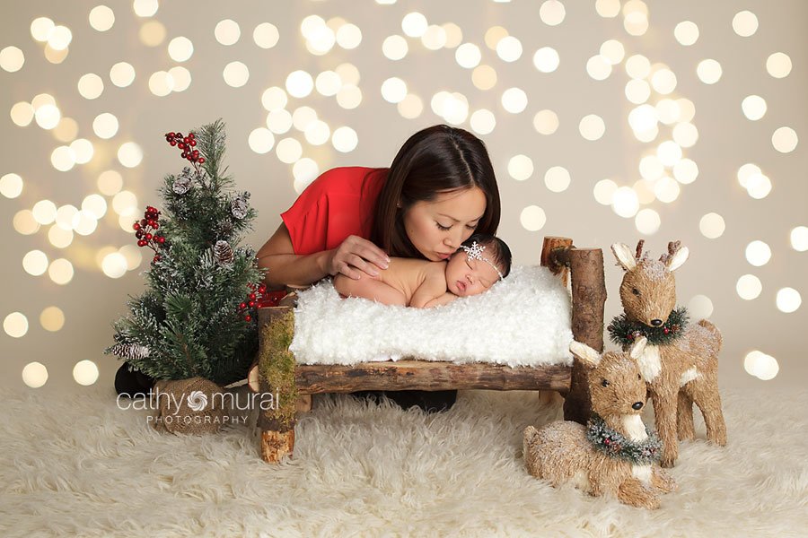 Beautiful mom kissing her newborn baby girl on the wooden bed during Christmas newborn portrait session. Edited with Christmas lights backdrop. Alhambra, San Gabriel Valley, CA Studio Newborn Photographer, Cathy Murai Photography 