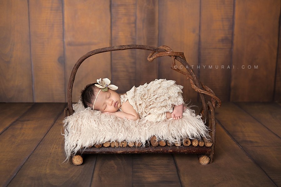 Sleeping Asian newborn baby girl wearing a cream lace romper and flower headband posing  on the wooden bed in front of the wooden backdrop. Captured by Cathy Murai Photography, Los Angeles Newborn Photographer, Alhambra Newborn Photographer, San Gabriel Newborn Baby Photographer, newborn props, newborn photography props.
