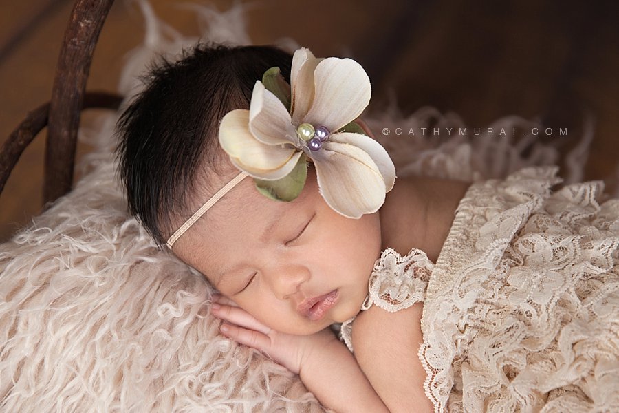 Close up of the sleeping Asian newborn baby girl wearing a cream lace romper and flower headband posing  on the wooden bed in front of the wooden backdrop. Captured by Cathy Murai Photography, Los Angeles Newborn Photographer, Alhambra Newborn Photographer, San Gabriel Newborn Baby Photographer, newborn props, newborn photography props.