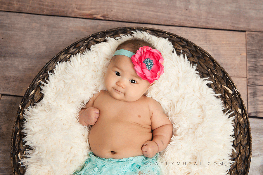 happy 3 month old baby wearing hot pink headband and light blue, aqua  lace romper laying on furry stuffing in the basket