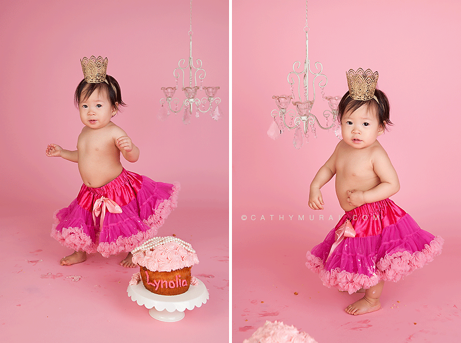 Adorable girl wearing a hot pink pettiskirt and gold crown standing behind the birthday giant cupcake dancing during first birthday portrait session , 1st birthday portrait session, pink theme, chandelier prop