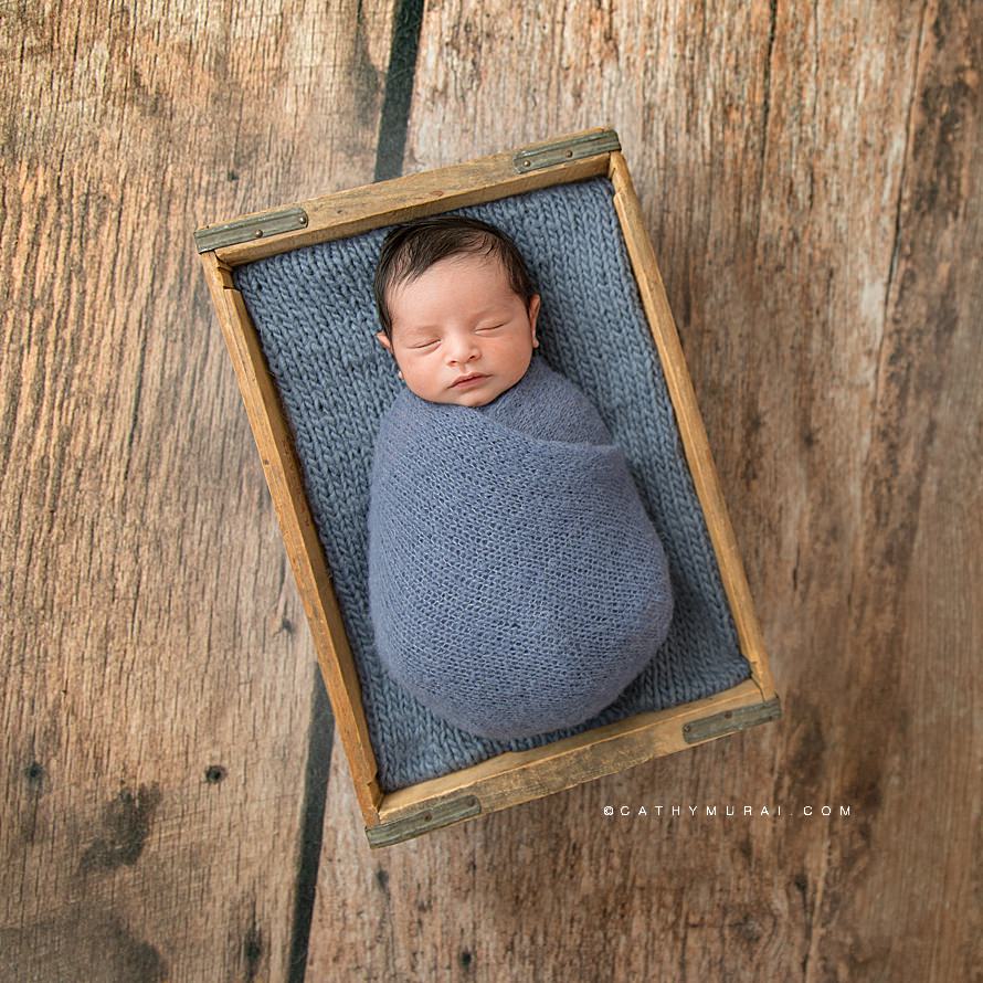 Newborn Session and Birth Announcement Photographs - Orange County Newborn Photographer - Cathy Murai Photography - photography studio based in Irvine, CA. Newborn baby boy swaddled in blue knitted wrap sleeping inside the vintage wooden box on the wood floor.  Square newborn photo. newborn photo in square