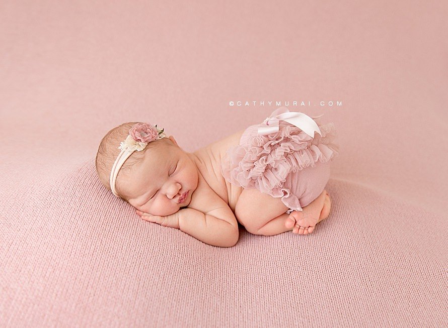 Cathy Murai Photography is a Irvine Newborn photographer based in Orange County, CA. This adorable newborn picture was captured in her Irvine Newborn Photography studio. Newborn baby girl wearing a blush pink headband and blush raffle diaper cover, posing Bum up pose, Tushy up pose while she was sleeping on the blush pink backdrop blanket during her newborn session in Irvine, CA.