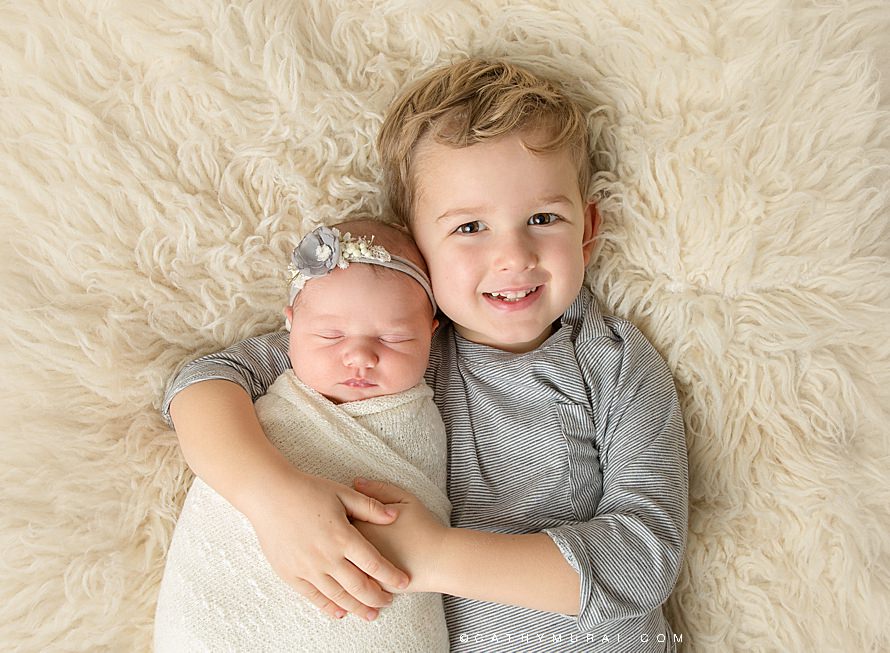 Cathy Murai Photography, Orange County Newborn photographer, Irvine Newborn Photographer, Sibling pose, Sibling relational pose, excited and proud older brother holding his sleeping little sister wrapped in a cream wrap and wearing a grey headband