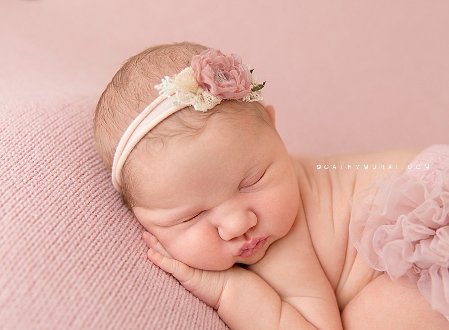Cathy Murai Photography is a Irvine Newborn photographer based in Orange County, CA. This adorable newborn picture was captured in her Irvine Newborn Photography studio. Newborn baby girl wearing a pink flower headband and pink blush raffle diaper cover, posing while she was sleeping on the blush pink backdrop blanket during her newborn session in Irvine, CA.