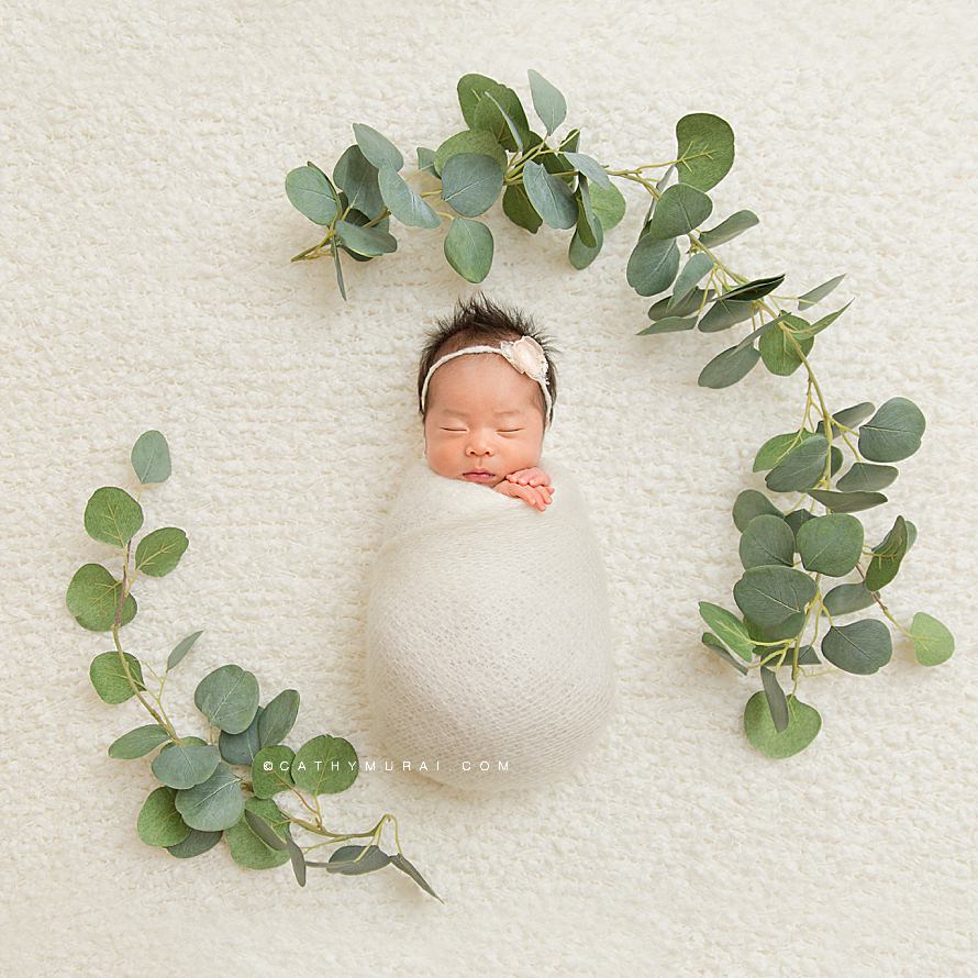 Natural Organic Newborn Photography Session by Cathy Murai Photography, Orange County Newborn and baby photographer in Irvine, a newborn baby girl wearing a cream headband and cream wrap is in a Wrapped Pose, newborn baby and Eucalyptus Leaf Garland prop
