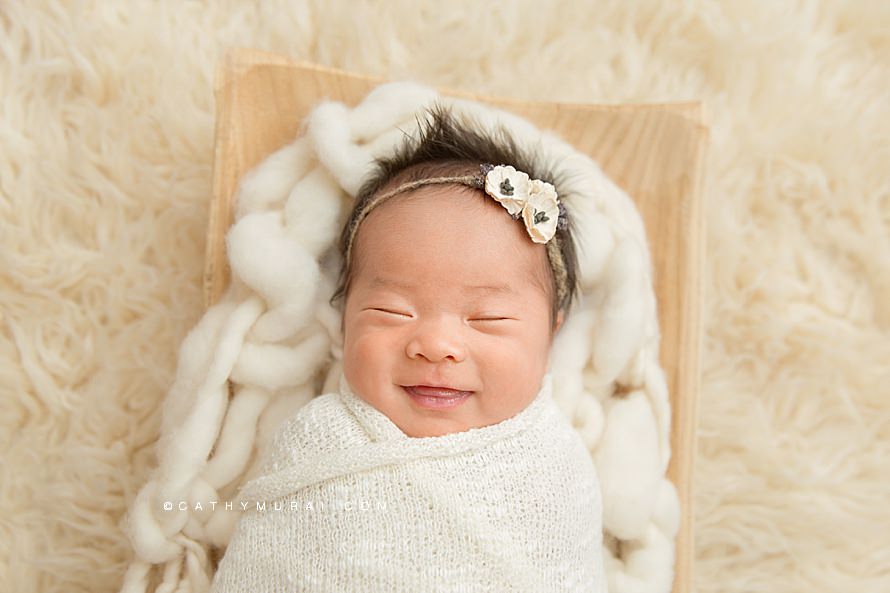 Natural Organic Newborn Photography by Cathy Murai Photography, Orange County Newborn and baby photographer in Irvine, a newborn baby girl wearing a cream headband is in simple wrapped pose on a wooden tray, smiling, closeup newborn image, smiling newborn photo, smiling newborn picture, smiling newborn baby portrait, a newborn baby with a big smile, newborn baby's adorable smile.jpg