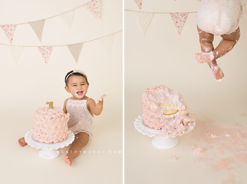 Getting messy at First Birthday Cake Smash Photography session with a vintage style using a pink cake, white cake stand, and a vintage banner, prop - captured by Cathy Murai Photography, a Irvine baby photographer
