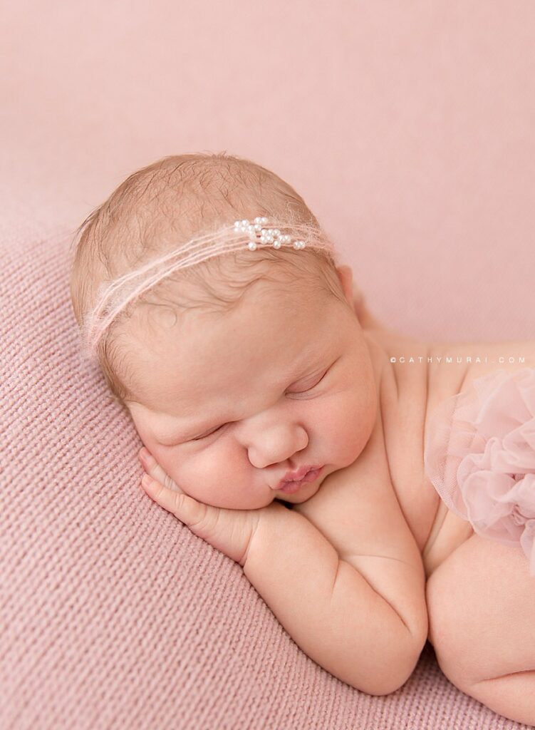 Cathy Murai Photography is a Irvine Newborn photographer based in Orange County, CA. This adorable newborn picture was captured in her Irvine Newborn Photography studio. Newborn baby girl wearing a blush pink and pearl headband and blush raffle diaper cover, posing Bum up pose, Tushy up pose while she was sleeping on the blush pink backdrop blanket during her newborn session in Irvine, CA. Newborn Close up shot.