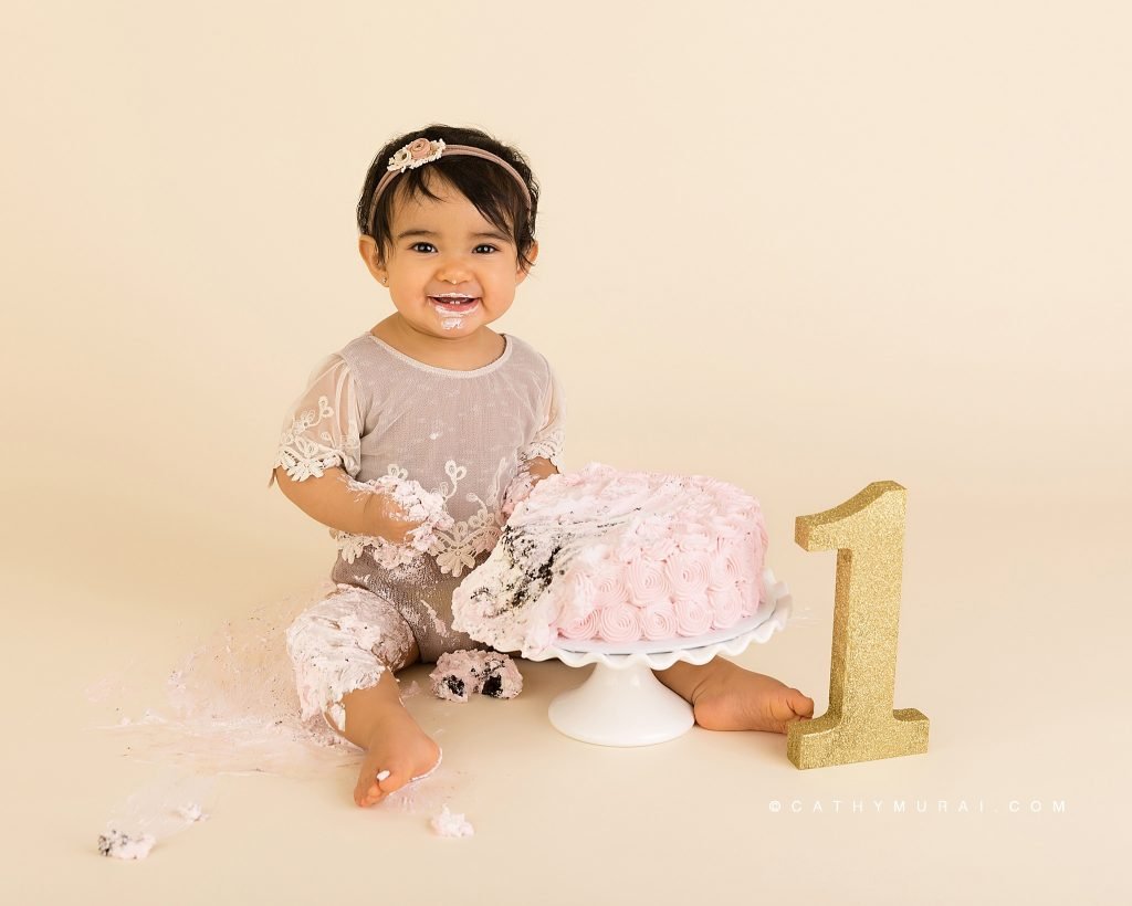 Getting messy at First Birthday Cake Smash Photography session with a vintage style using a pink cake, white cake stand, and a gold number one prop by Cathy Murai Photography, a Irvine baby photographer