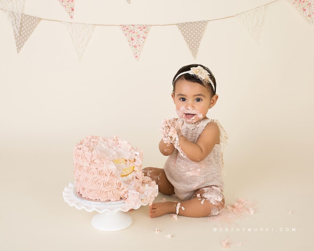 Getting messy during First Birthday Cake Smash Photography session with a vintage style using a pink cake, white cake stand, and a vintage banner, captured by Cathy Murai Photography, a Irvine baby photographer