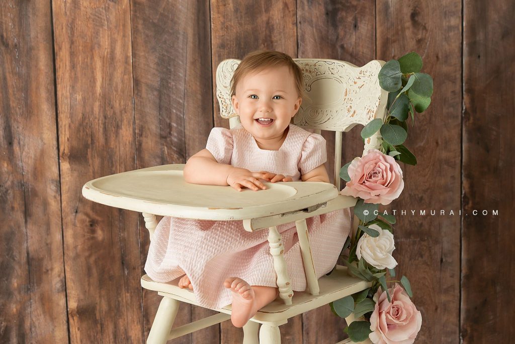 happy first birthday girl on a white vintage high chair / cream vintage high chair decorated with pink flowers during her first birthday photoshoot with Cathy Murai Photography, a Irvine baby photographer