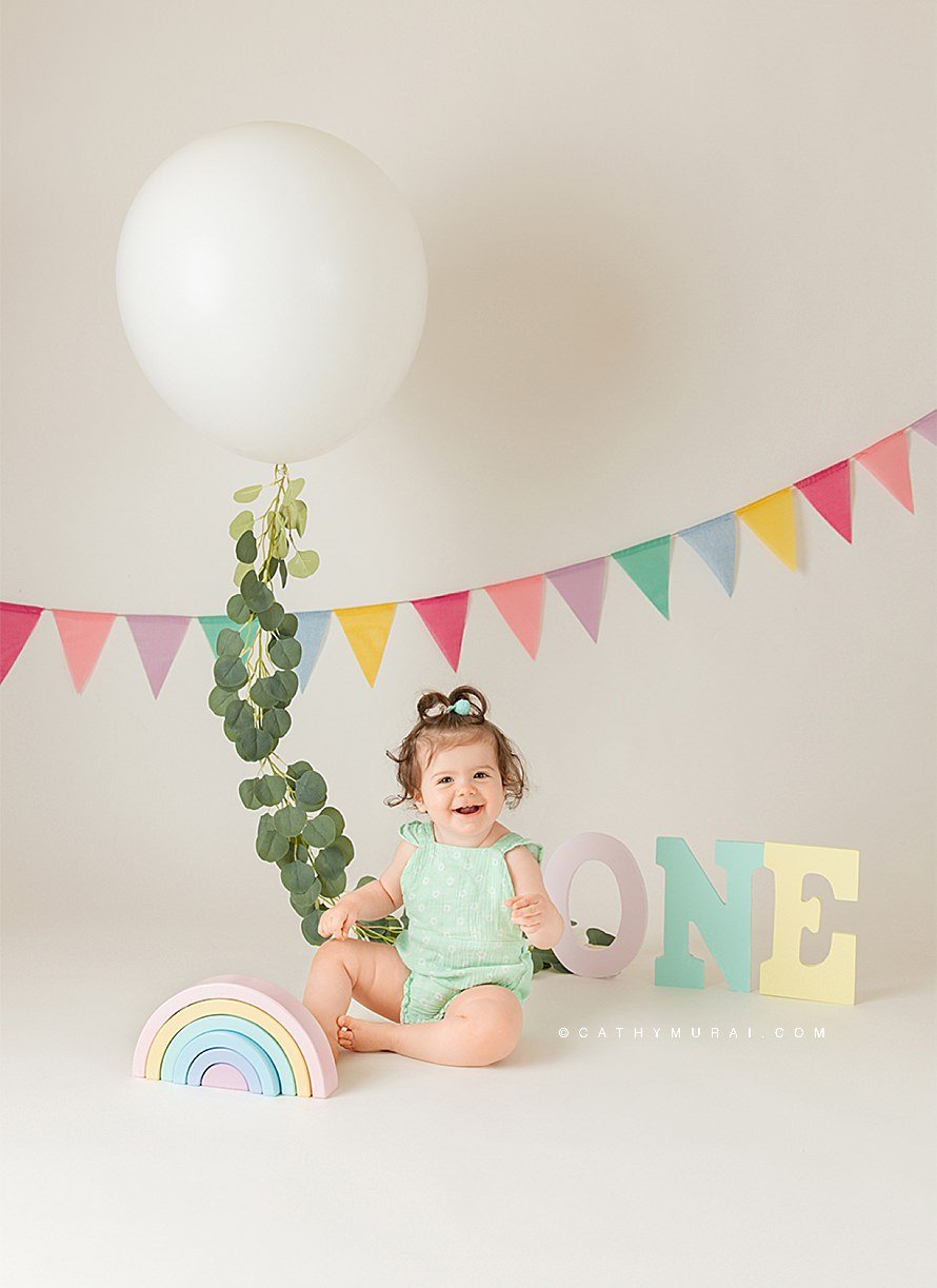 Pastel rainbow themed first birthday photo session using props including a rainbow wooden toy, o.n.e. letters in the pastel color, pastel banner, and a pastel outfit for the first birthday girl.