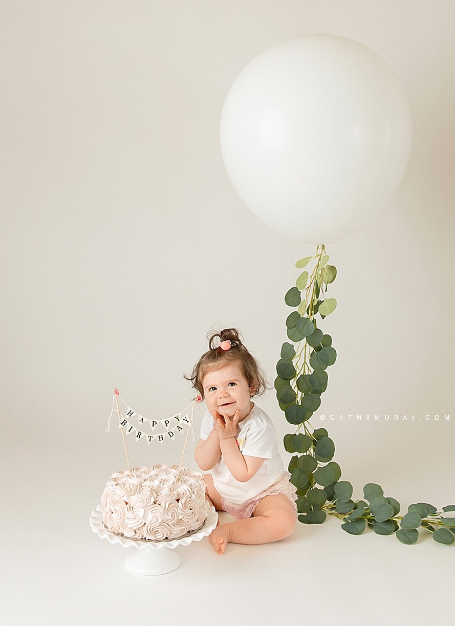 Simple white first birthday smash cake session using one big white balloon  with artificial garland and a birthday cake with a happy birthday banner.