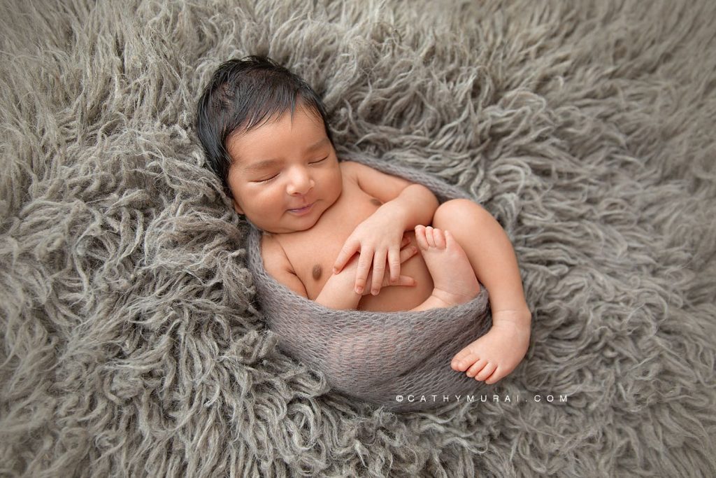 Newborn baby boy with a grey wrap smiled at his newborn photoshoot Covid-19 with Cathy Murai Photography