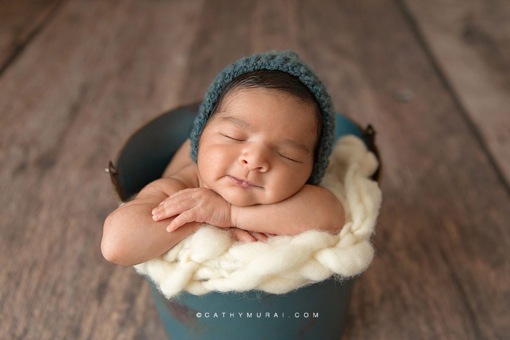 Indian newborn baby boy wearing a teal knit hat posing in a teal bucket prop during his newborn photoshoot Covid-19 with Cathy Murai Photography, orange county newborn photographer, in Irvine, CA