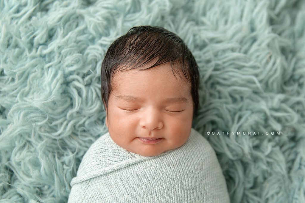 Newborn baby boy swaddled in a teal wrap laying on a furry, teal background, smiling while sleeping, during his newborn photo session with Cathy Murai Photography, orange county newborn photographer, in her studio in Irvine