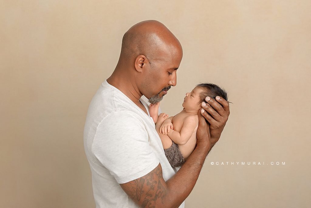Father holds and looks at his newborn baby boy who is sleeping while wearing a brown, knit diaper cover in front of a beige backdrop. Newborn with father photo taken by Cathy Murai Photography, an OC Newborn Photographer in Irvine, CA