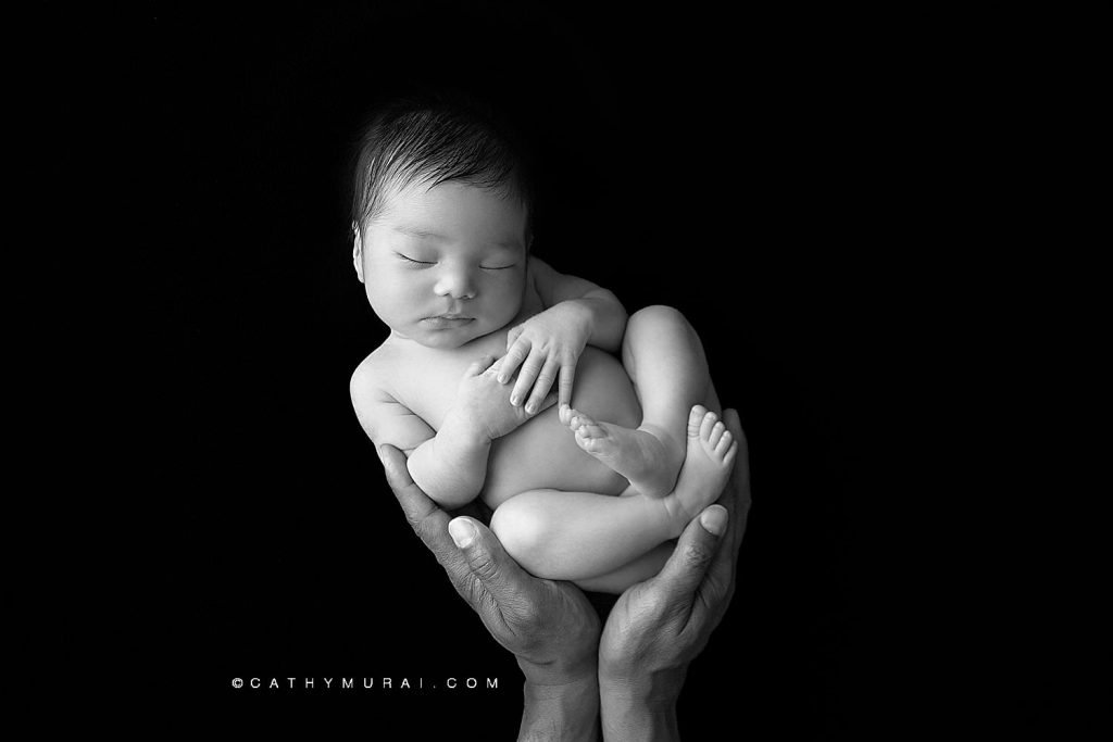 Newborn baby boy with dark hair sleeps while being held in father's hands in front of a black background.