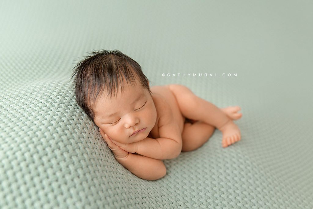 Newborn baby boy with dark hair sleeps on a teal colored blanket while leaning on hands.