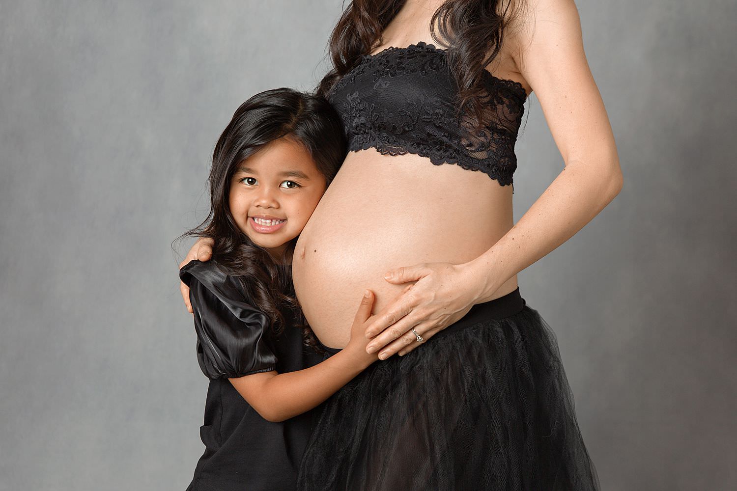 Mother-Daughter Maternity Photoshoot by Cathy Murai Photography, Orange County Maternity Photographer. Pregnant mother and her daughter, both in black dresses and holding pregnant belly, in front of gray backdrop during maternity photoshoot