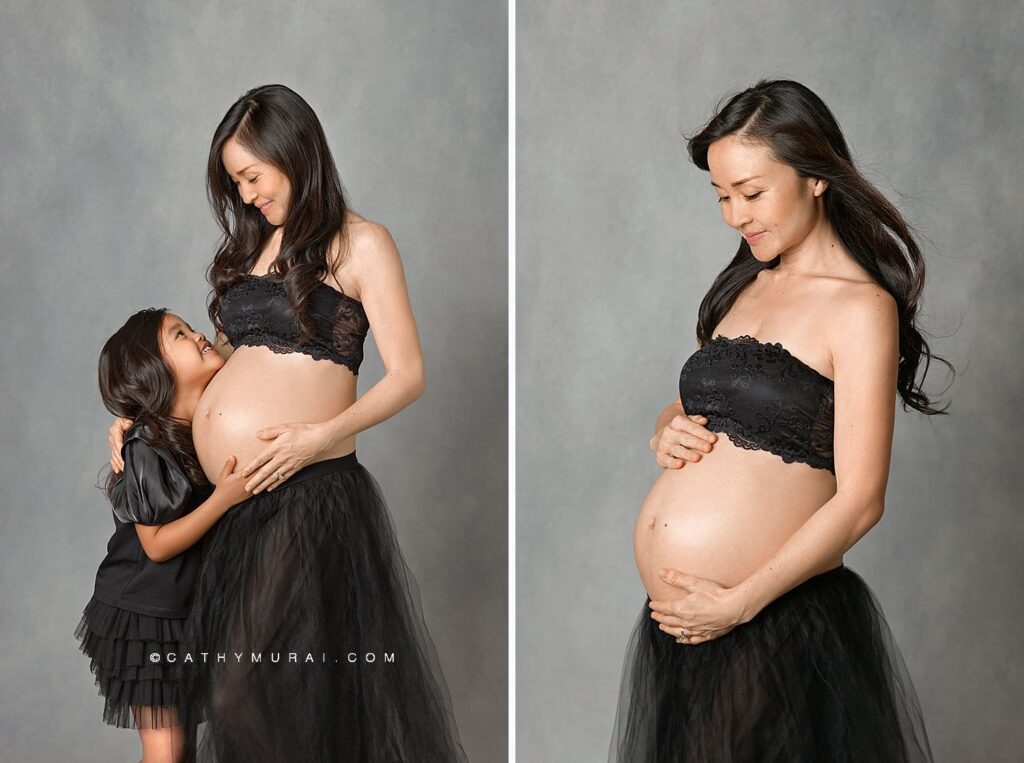 Pregnant mother and her daughter, both in black dresses, holding pregnant belly in front of gray backdrop during maternity photoshoot with Cathy Murai Photography