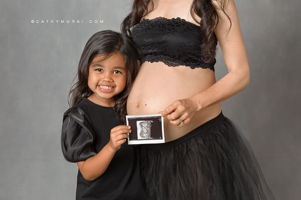 Mother-Daughter Maternity Photoshoot by Cathy Murai Photography, Orange County Maternity Photographer. Pregnant mother and her daughter, both in black dresses, holding ultrasound image in front of gray backdrop during maternity photoshoot