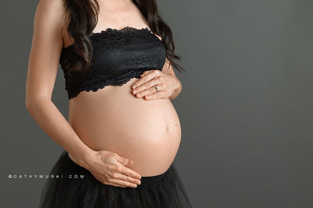 Pregnant mother in black tulle skirt and black lace bandeau holding belly in front of gray backdrop during maternity photoshoot with Cathy Murai Photography