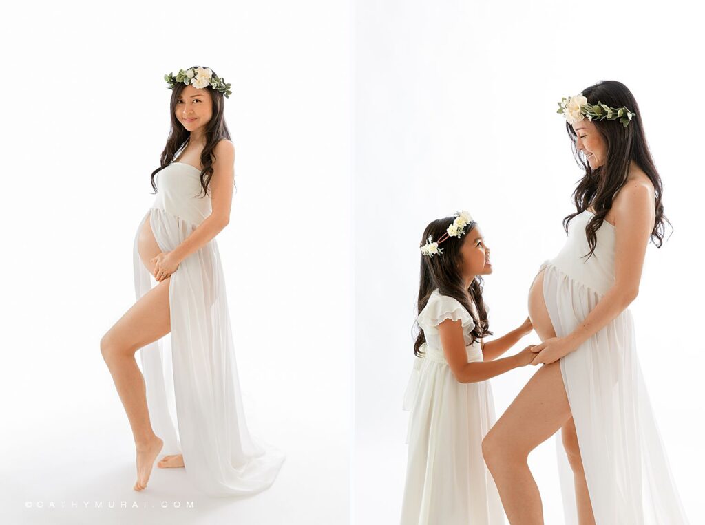 Mother-Daughter Maternity Photoshoot by Cathy Murai Photography, Orange County Maternity Photographer.  Pregnant mother and her daughter, both in white dresses and flower crowns, looking at each other in front of white backdrop during maternity photoshoot