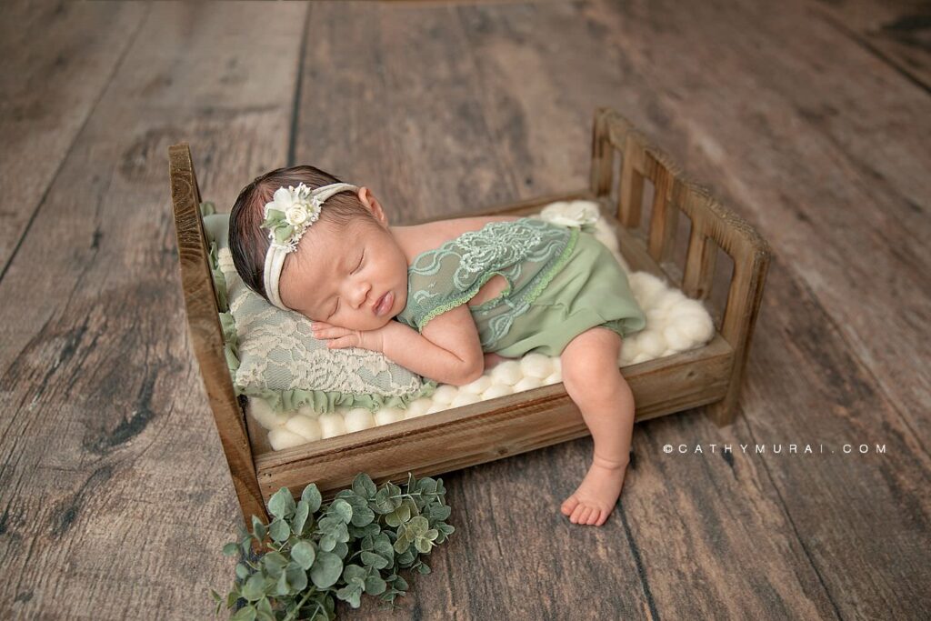 Newborn baby girl wearing a floral headband and sage green romper sleeps on a miniature bed with a soft white blanket and sage green pillow.