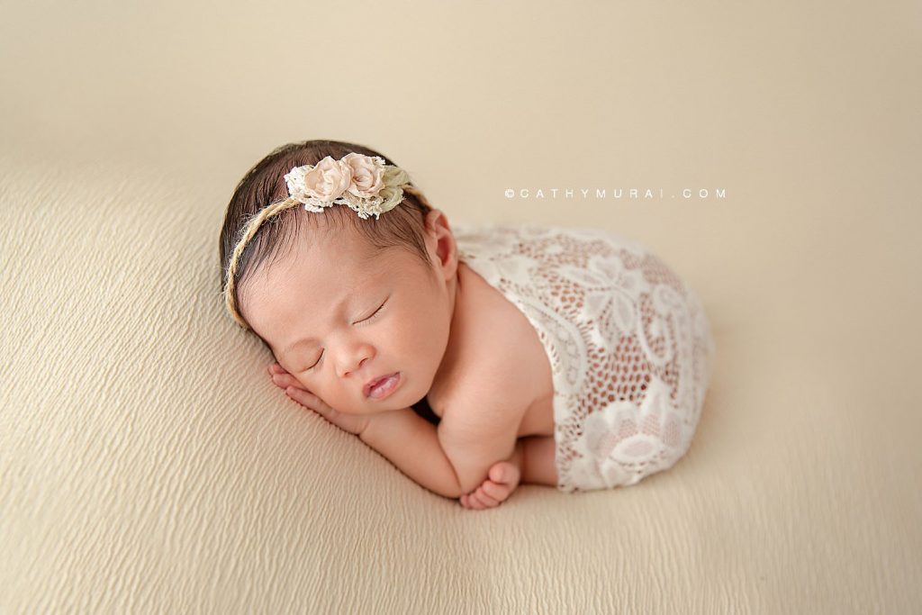 Newborn baby girl wearing a floral headband and white lace swaddle sleeps on a cream-colored blanket.