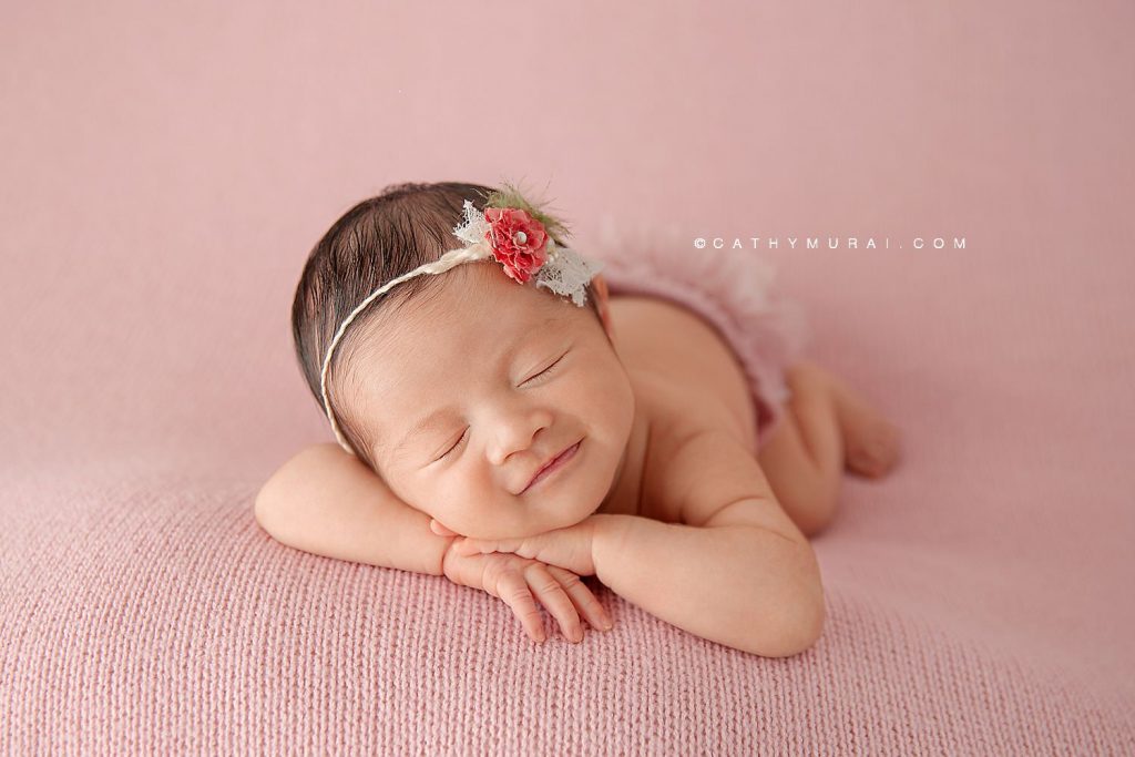Newborn baby girl wearing a floral headband and pink diaper cover sleeps on a pink blanket with a soft smile.