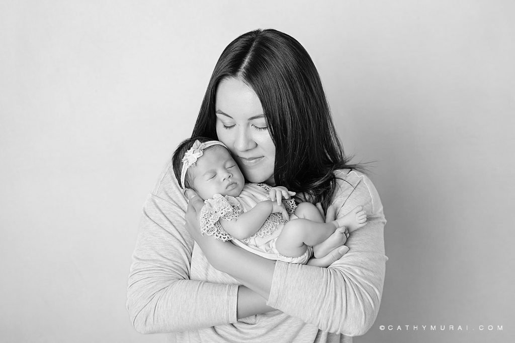 black and white image of a mom holding her sleeping newborn baby girl who wears a lace onesie and floral headband.