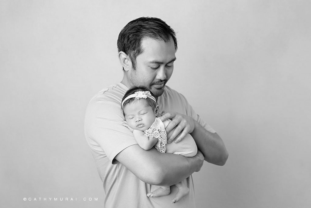 black and white mage of a dad holding his sleeping newborn baby girl who wears a lace onesie and floral headband.