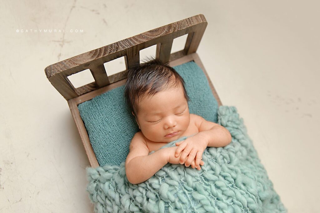 A newborn baby boy posing and sleeping on a wooden bed with teal blanket while Cathy Murai Photography captured his newborn photos.