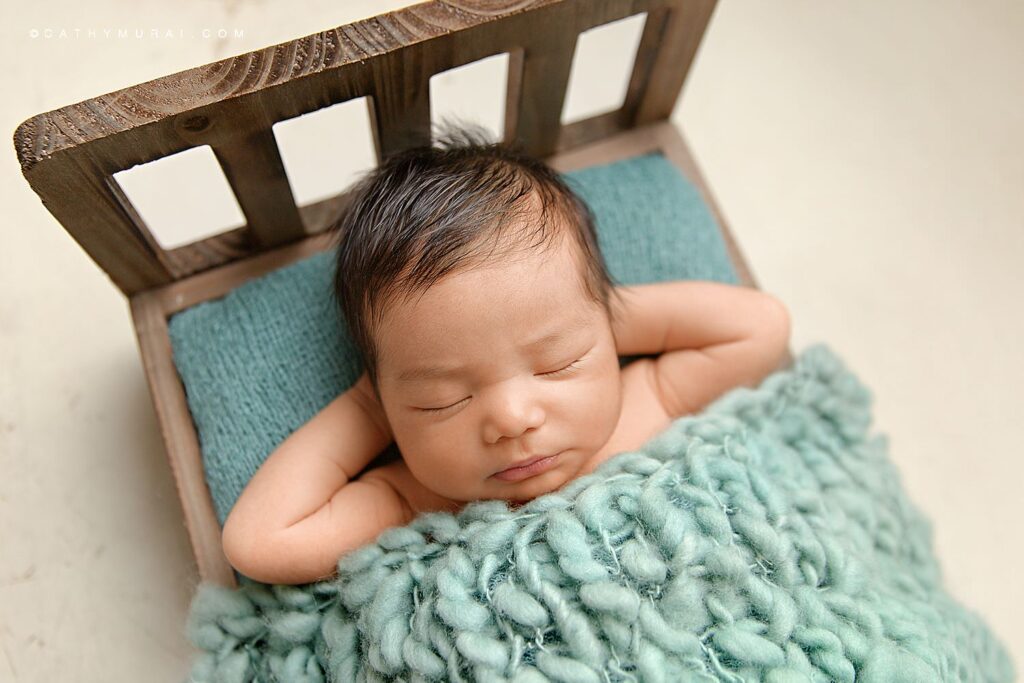 A newborn baby boy sleeping and posing on a wooden bed with teal blanket while Cathy Murai Photography captured his newborn photos.