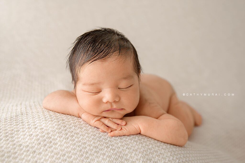 A newborn baby boy in chin on hands pose while sleeping on a cream and beige blanket during his newborn photo session in studio in Irvine, CA
